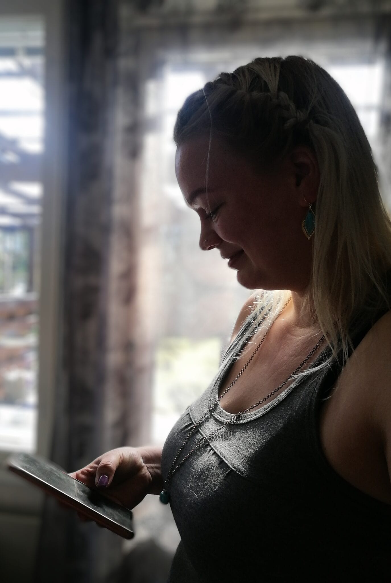 ikkuna_taustalla_varjoinen_kuva_nainen_seisoo_puhelin_karessa_hymyillen_katse_kaannetty_puhelimeen