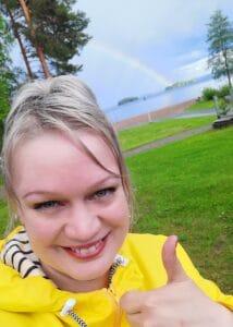 strateginen_kumppani_verkko_valkyrie nainen hymyilee kameraa kohti karsellen peukku pystyssä taustalla sateenkaari ja järvimaisema