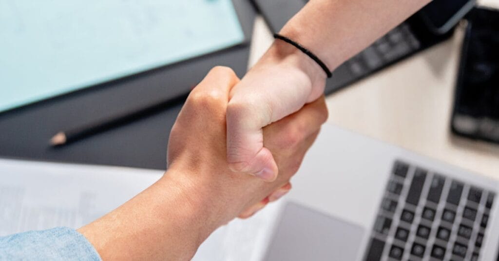 Two people shaking hands over laptops and documents in an office, symbolizing a business deal.