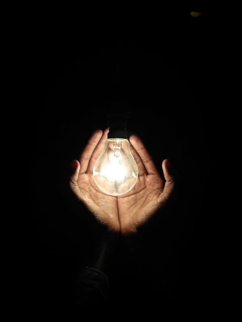 A pair of hands gently cradles a glowing light bulb, illuminating the darkness.