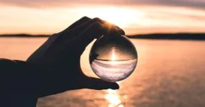 A hand holds a crystal ball reflecting a serene sunset over calm ocean waters.