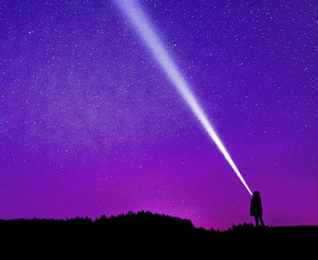 Silhouette of a person shining a flashlight under a starry violet night sky, creating a dramatic scene.